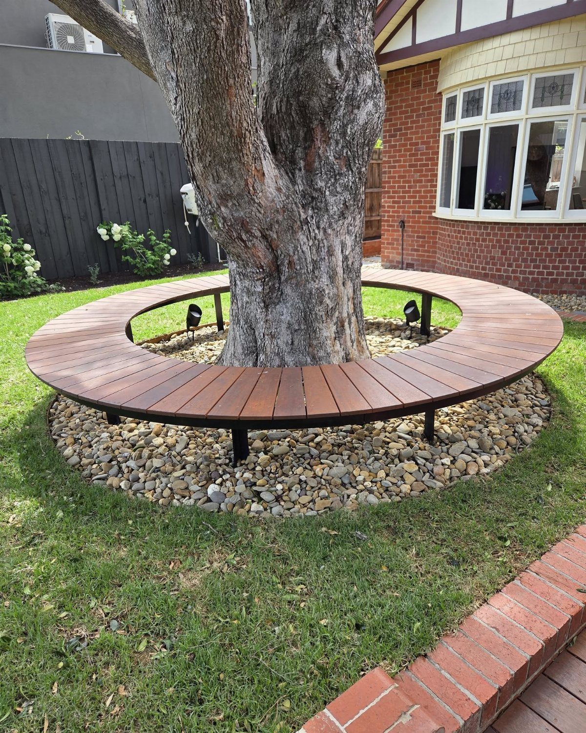 10 Around a Tree Ideas for Better Outdoor Spots 1 A circular wooden bench encircling a large tree trunk on a bed of stones.