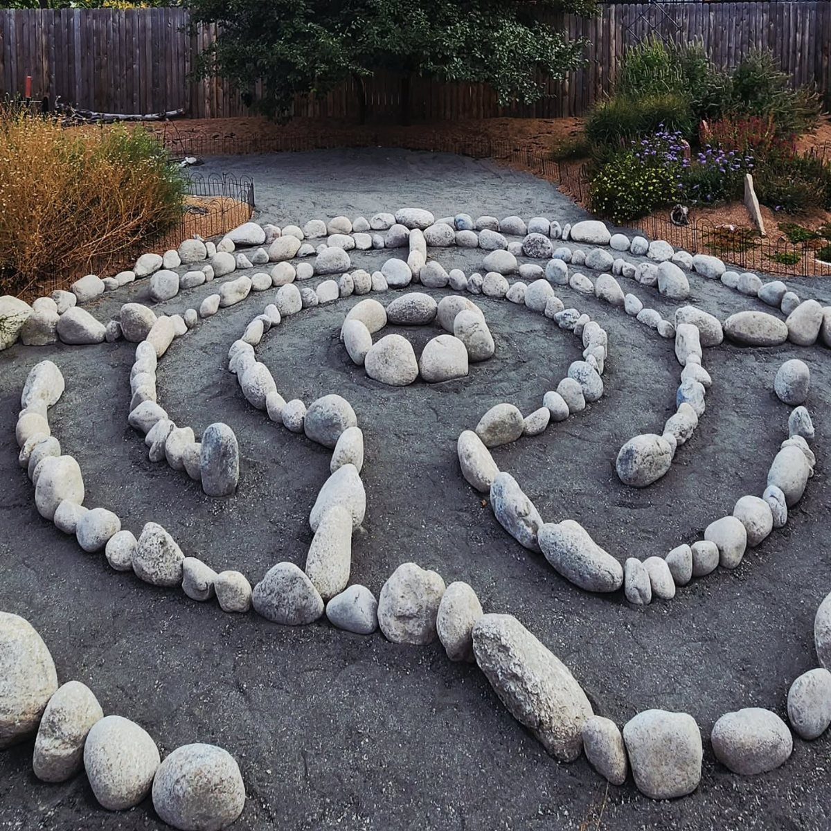 18 Big Backyard Garden Ideas for Every Budget 14 Light grey stones arranged in a winding labyrinth pattern on dark soil in a garden.