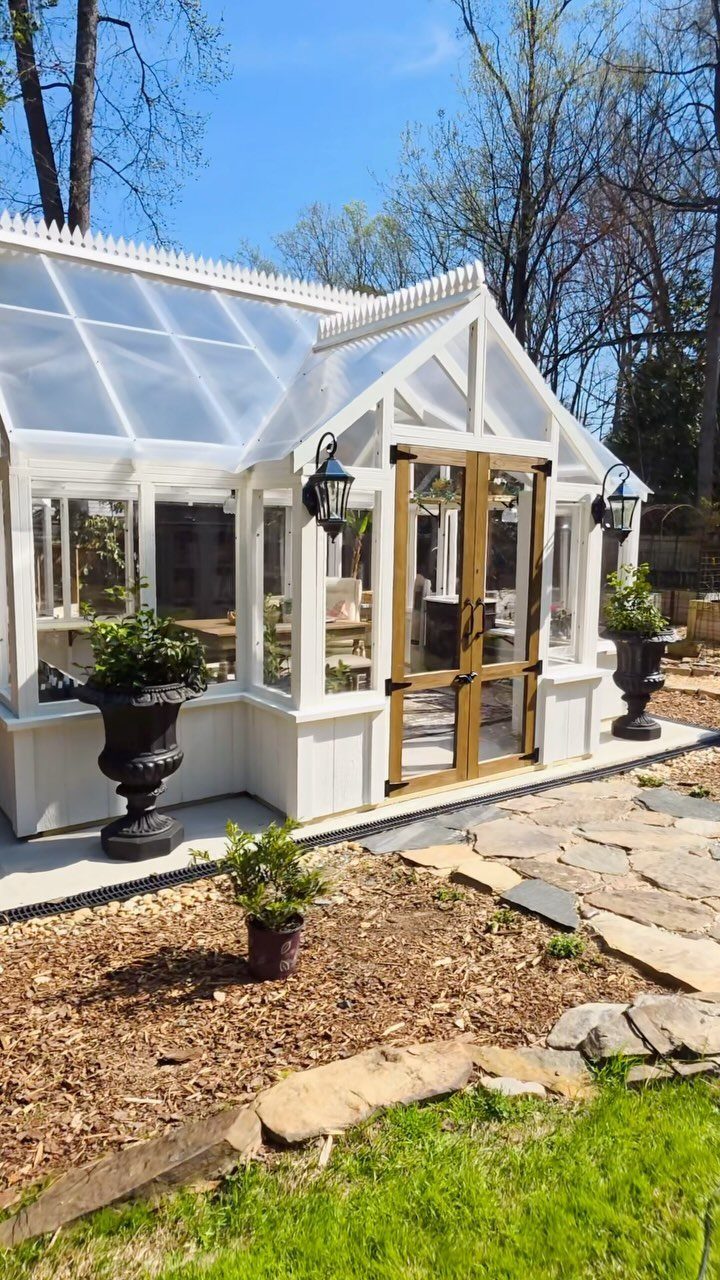 18 Big Backyard Garden Ideas for Every Budget 13 White wooden greenhouse with glass windows and wooden doors situated in a landscaped backyard garden.
