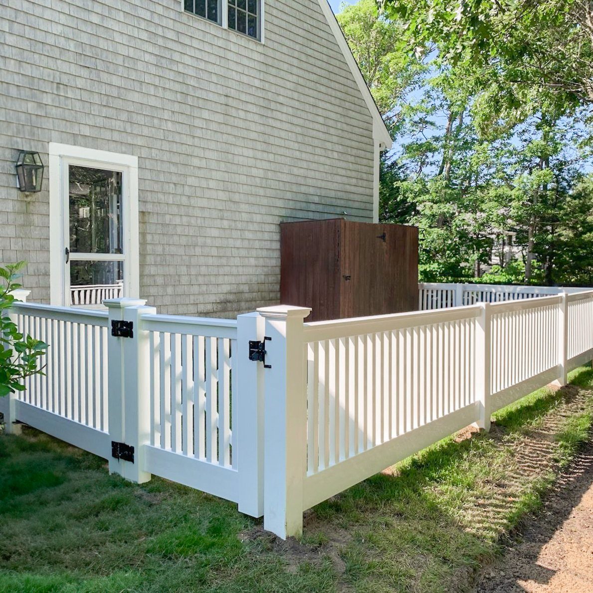 10 Cape Cod Front Yard Landscaping Ideas for Classic Charm 3 White picket fence with decorative gate panels bordering a green lawn next to a shingled house.