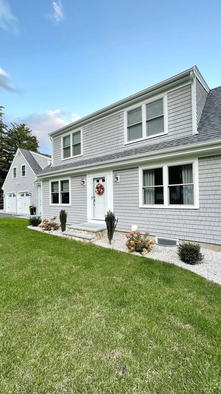 10 Cape Cod Front Yard Landscaping Ideas for Classic Charm 10 A grey shingle house with white trim, small shrubs, and a green lawn in the front yard.
