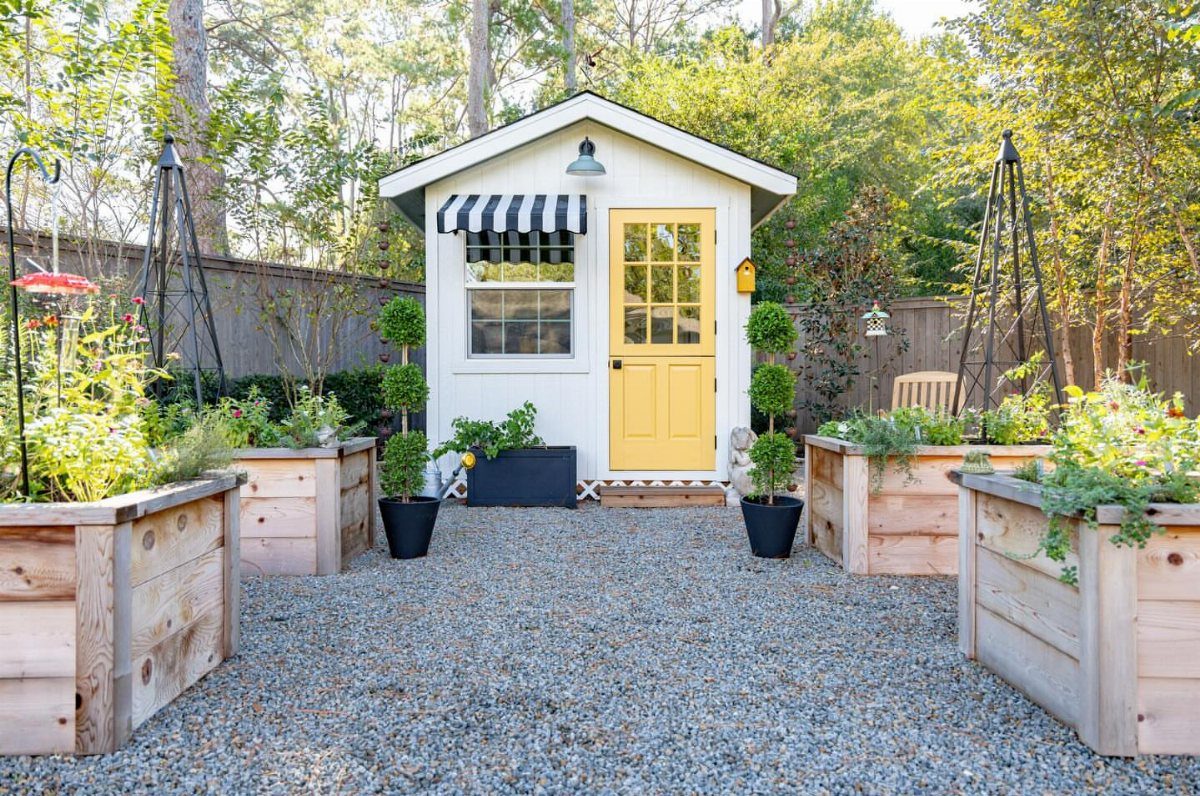 White cottage shed with a yellow door and striped awning centered between wooden garden planters.