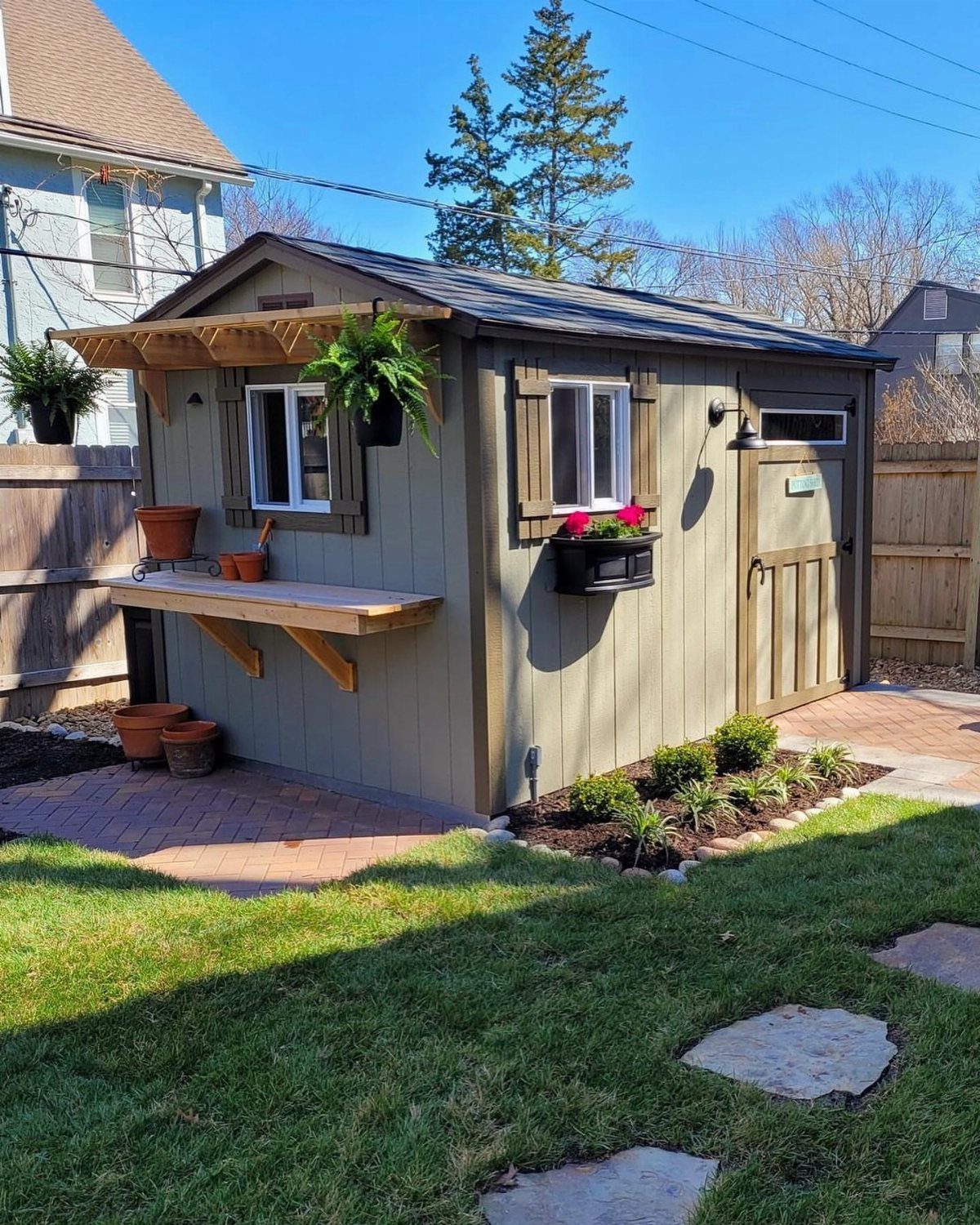 Small olive-green wooden garden shed with brown shutters and plants next to a brick patio.