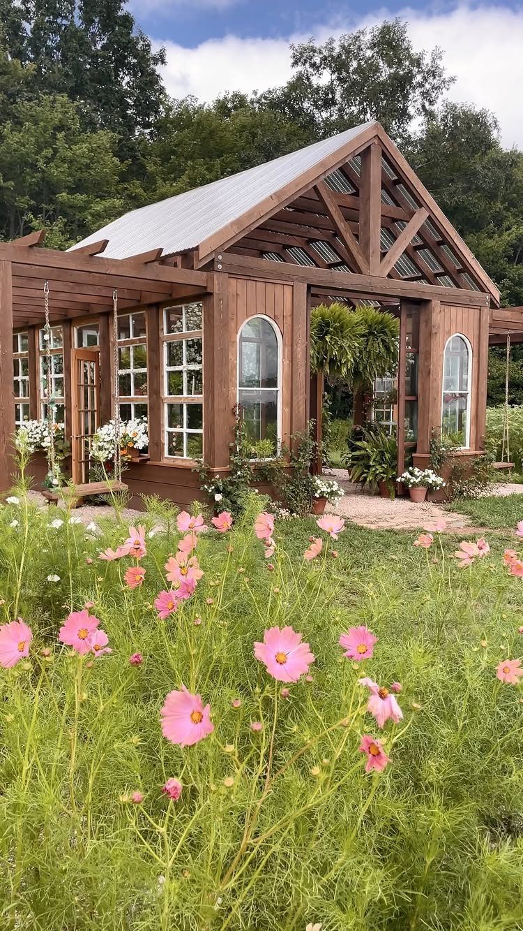 Wooden cottage garden structure with arched windows and an open front surrounded by pink cosmos flowers.