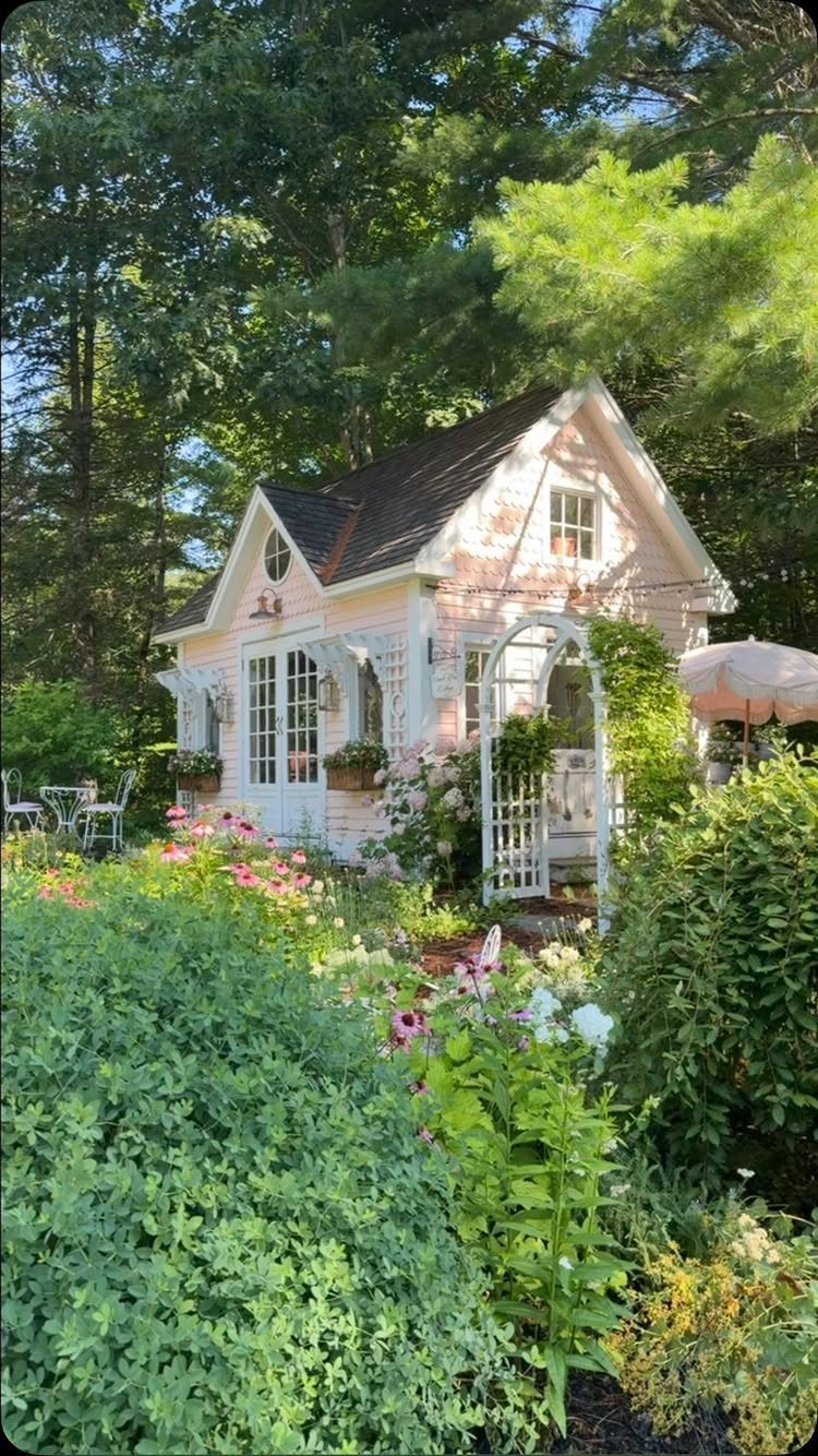 A light pink cottage garden shed surrounded by lush green plants and colorful flowers.