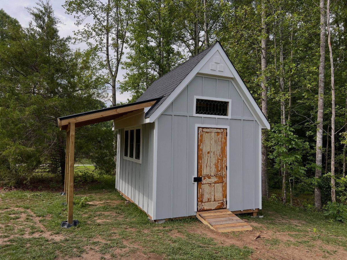 A light grey cottage-style wooden shed with white trim and a natural wood door in a grassy clearing.