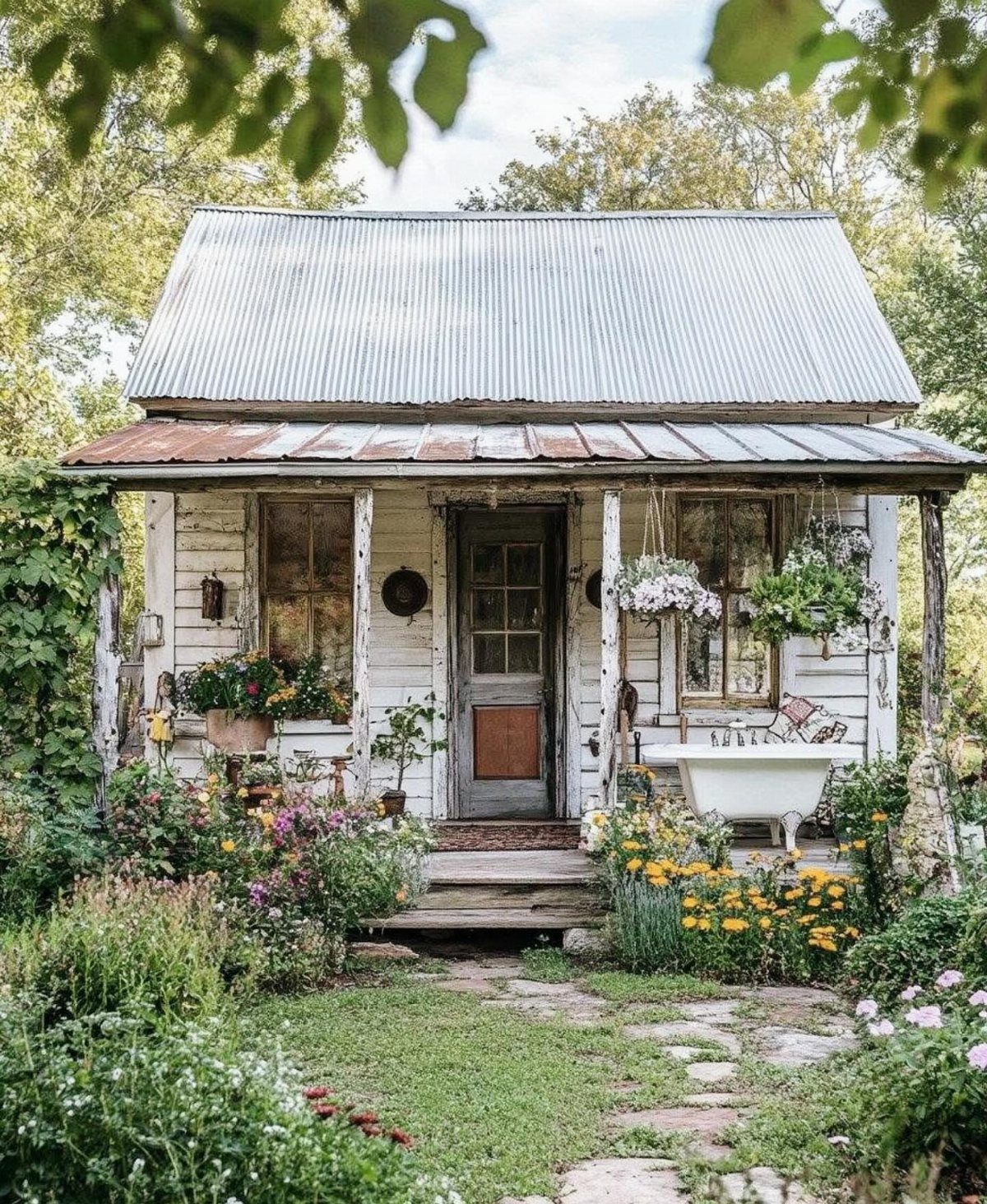 A weathered white wooden cottage shed surrounded by a blooming flower garden and a clawfoot bathtub.