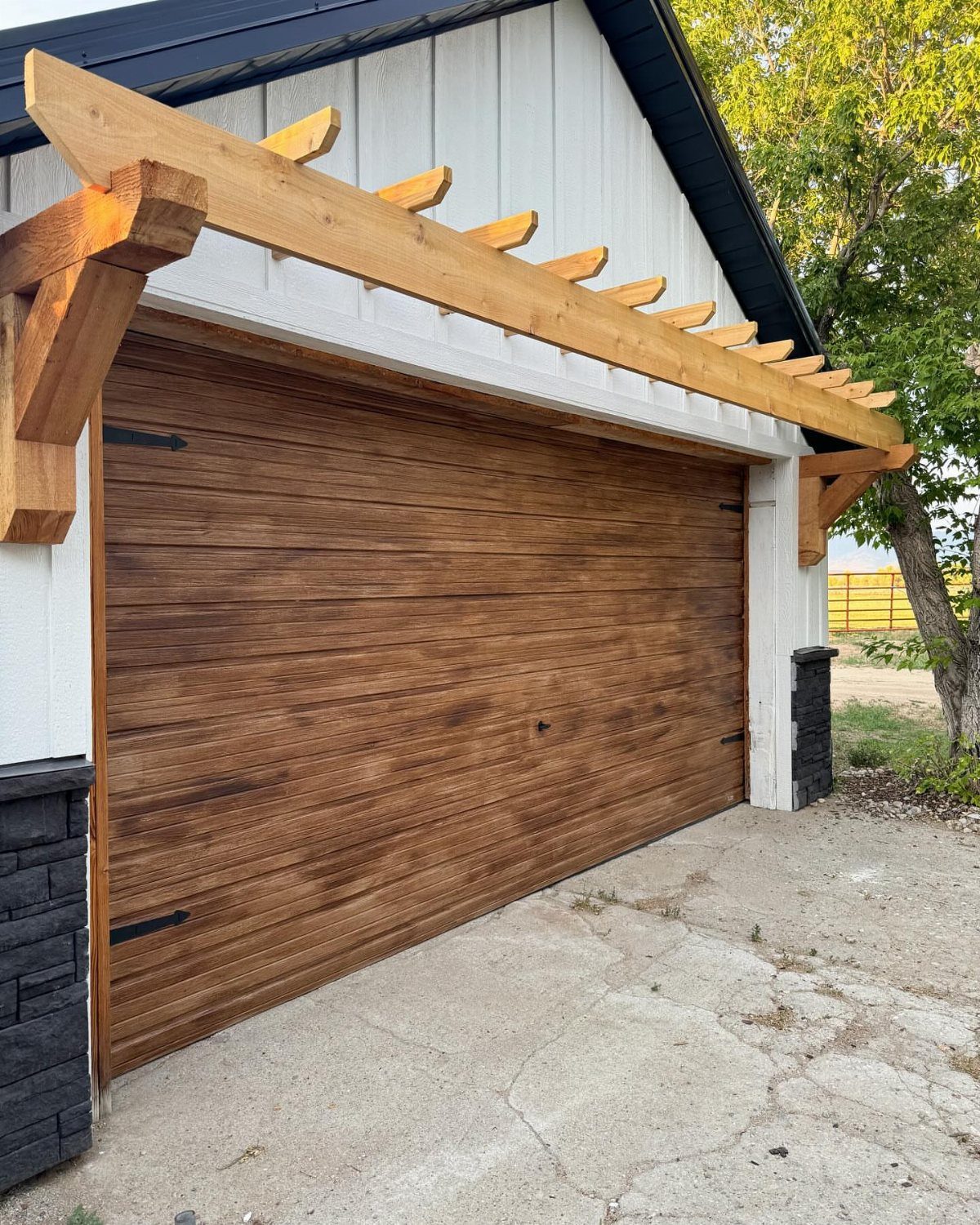 Craftsman Style Garage Doors: 8 Ideas from Carriage House to Modern Grey 1 A garage door with a brown faux-wood finish under a wooden pergola structure.
