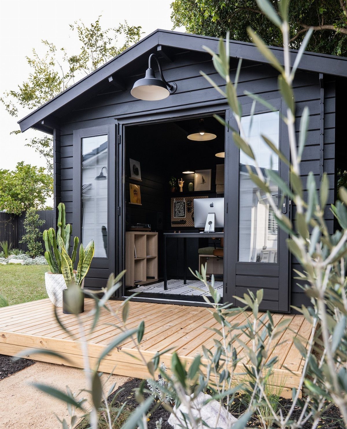 Black wooden shed with open doors revealing a workspace, sitting on a light wood deck.