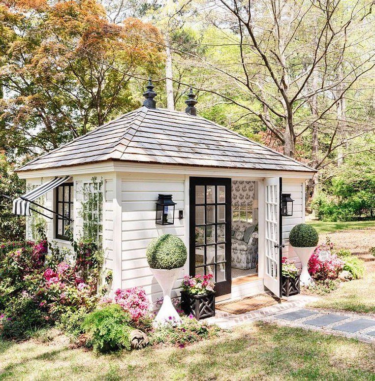 White wooden garden shed with black metal lanterns and glass doors surrounded by pink flowers.