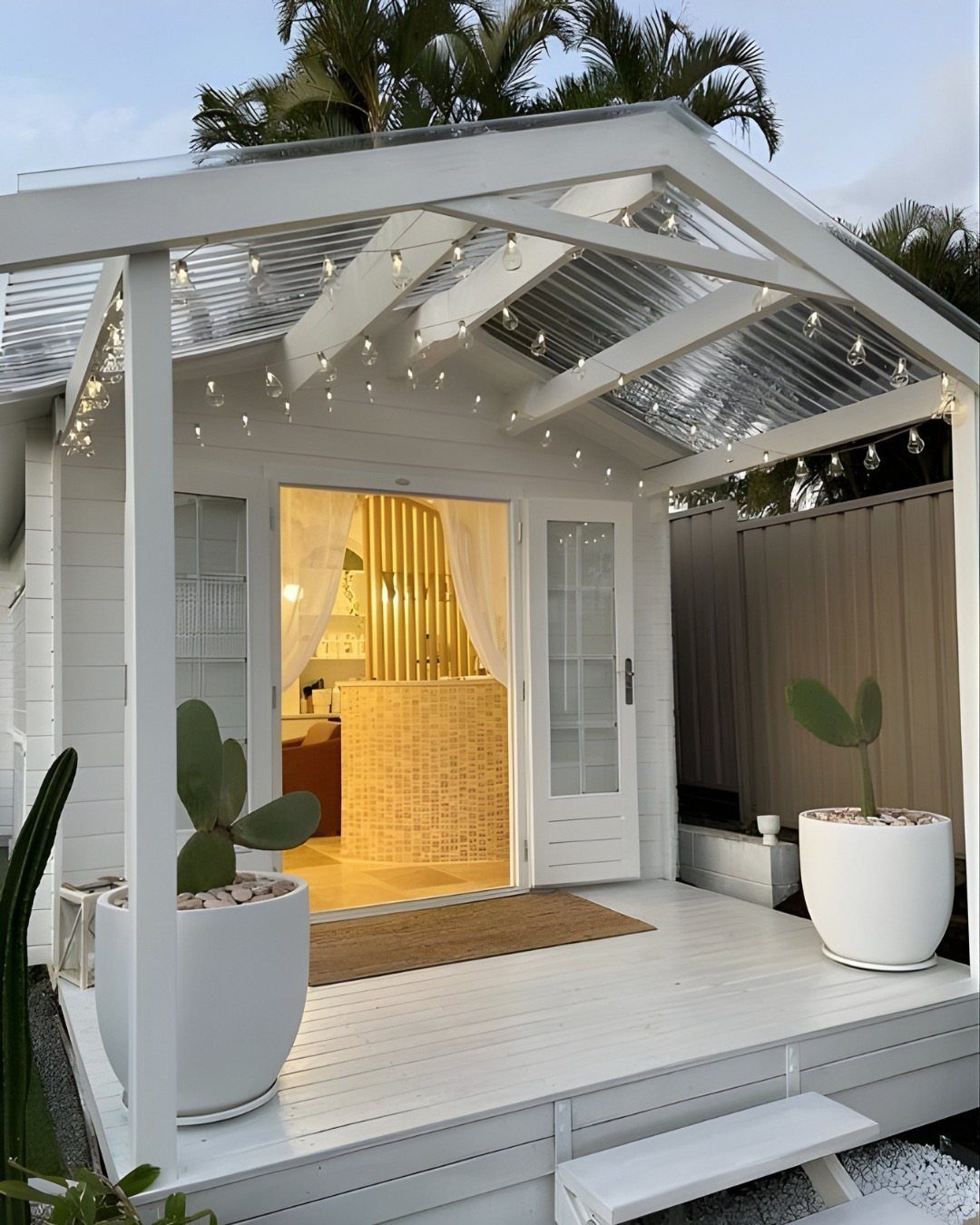 White wooden shed with a corrugated roof, string lights, and large white planters containing cacti on a deck.
