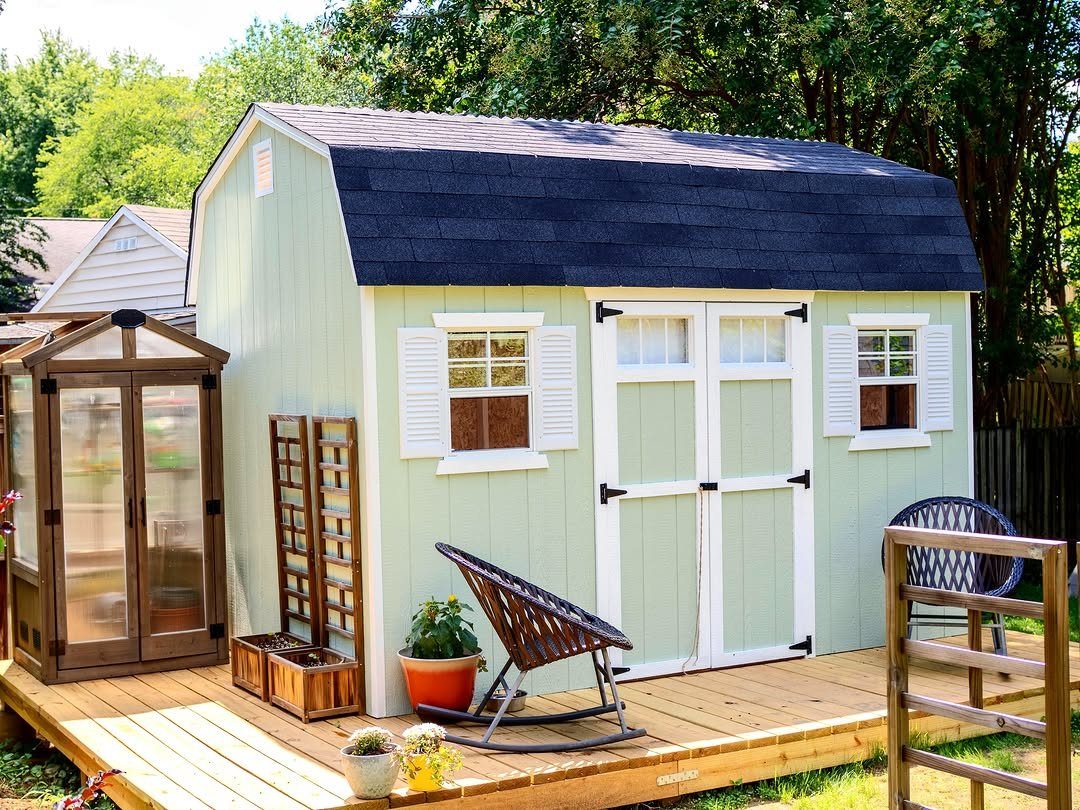 Light green garden shed with white shutters situated on a raised wooden deck with plants.