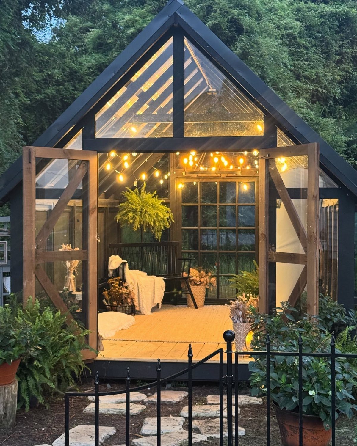 Glass garden shed with warm string lights, wooden floors, and white textiles inside a lush garden.