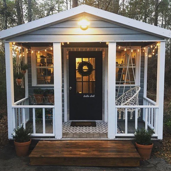 Small grey wooden shed with white trim, string lights, and a hanging chair on a porch.