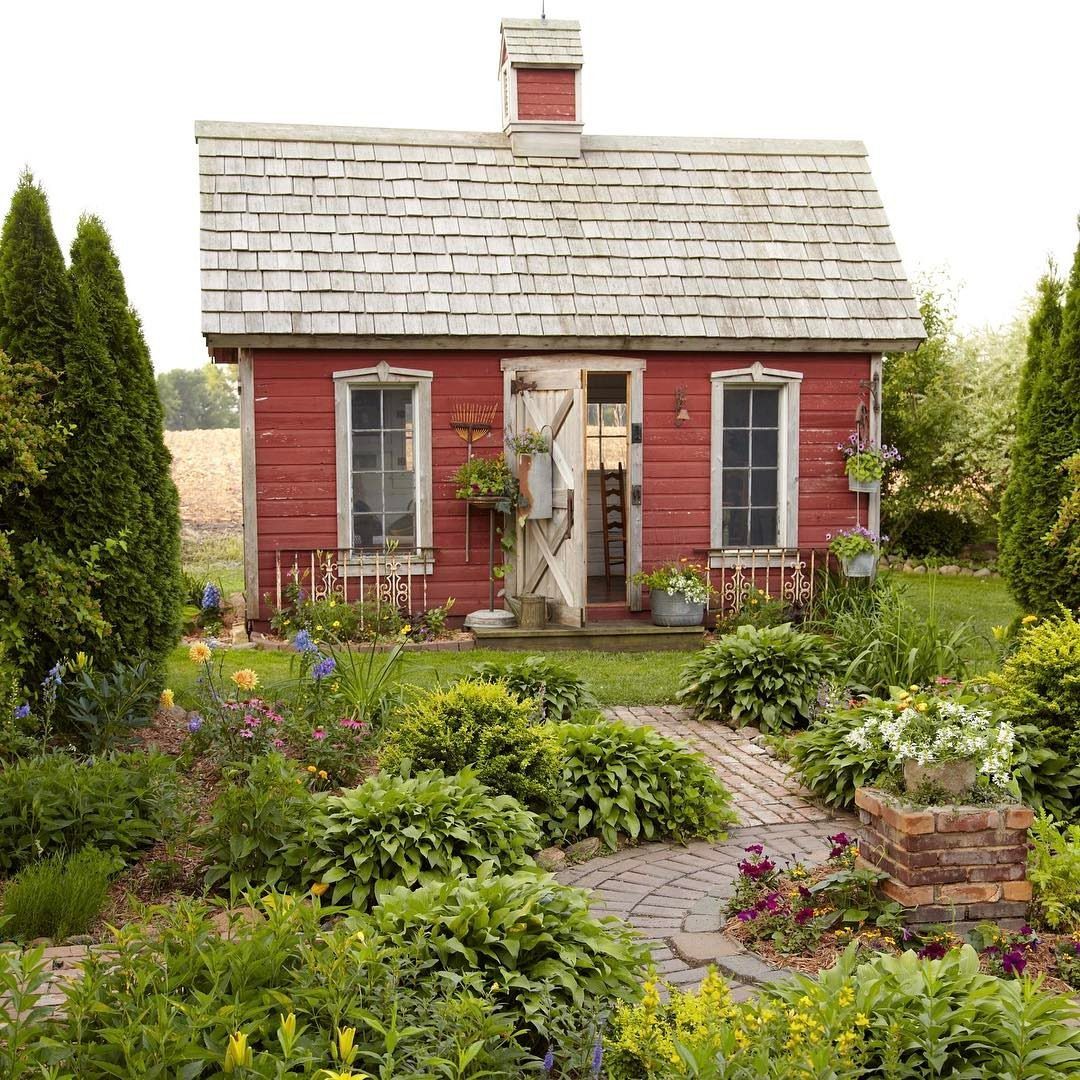 Red vintage wooden shed surrounded by a lush garden with a brick pathway and colorful flowers.
