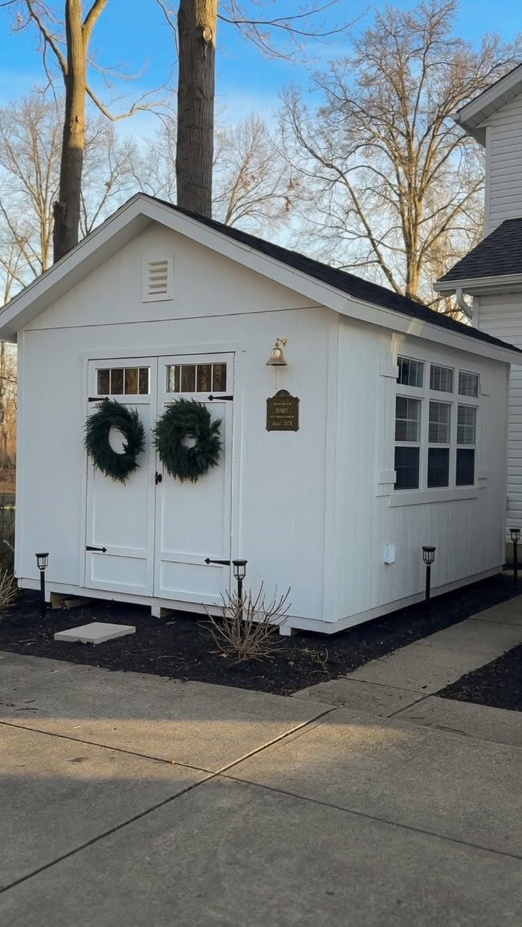 White wooden shed with double doors decorated with two green wreaths under a blue sky.