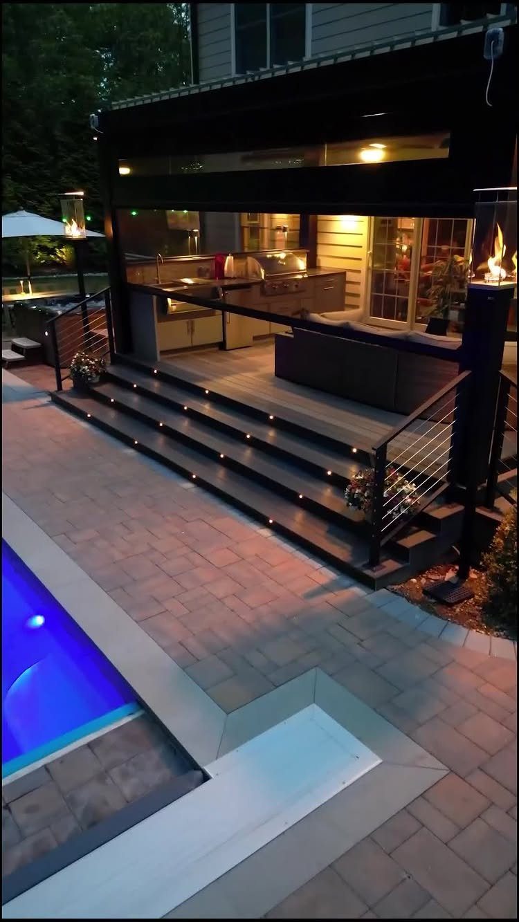 17 Deck Covering Ideas for Shade, Style & Rain Shelter 2 Modern outdoor kitchen and lounge area on a lit wooden deck next to a pool.