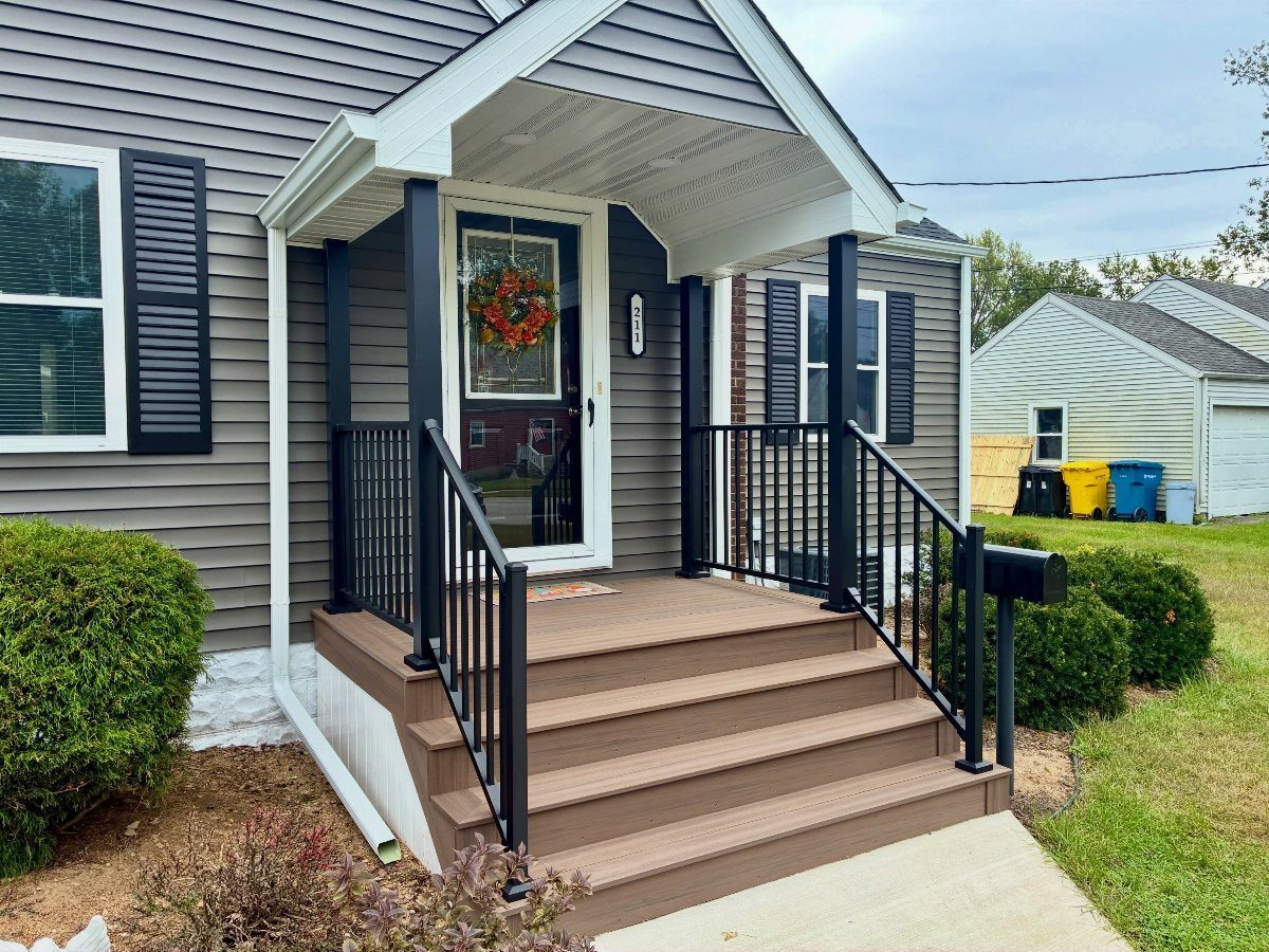 Deck on Front of House: 10 Ideas for Welcoming Style 9 A small grey house featuring a brown wooden porch with black metal railings and steps.