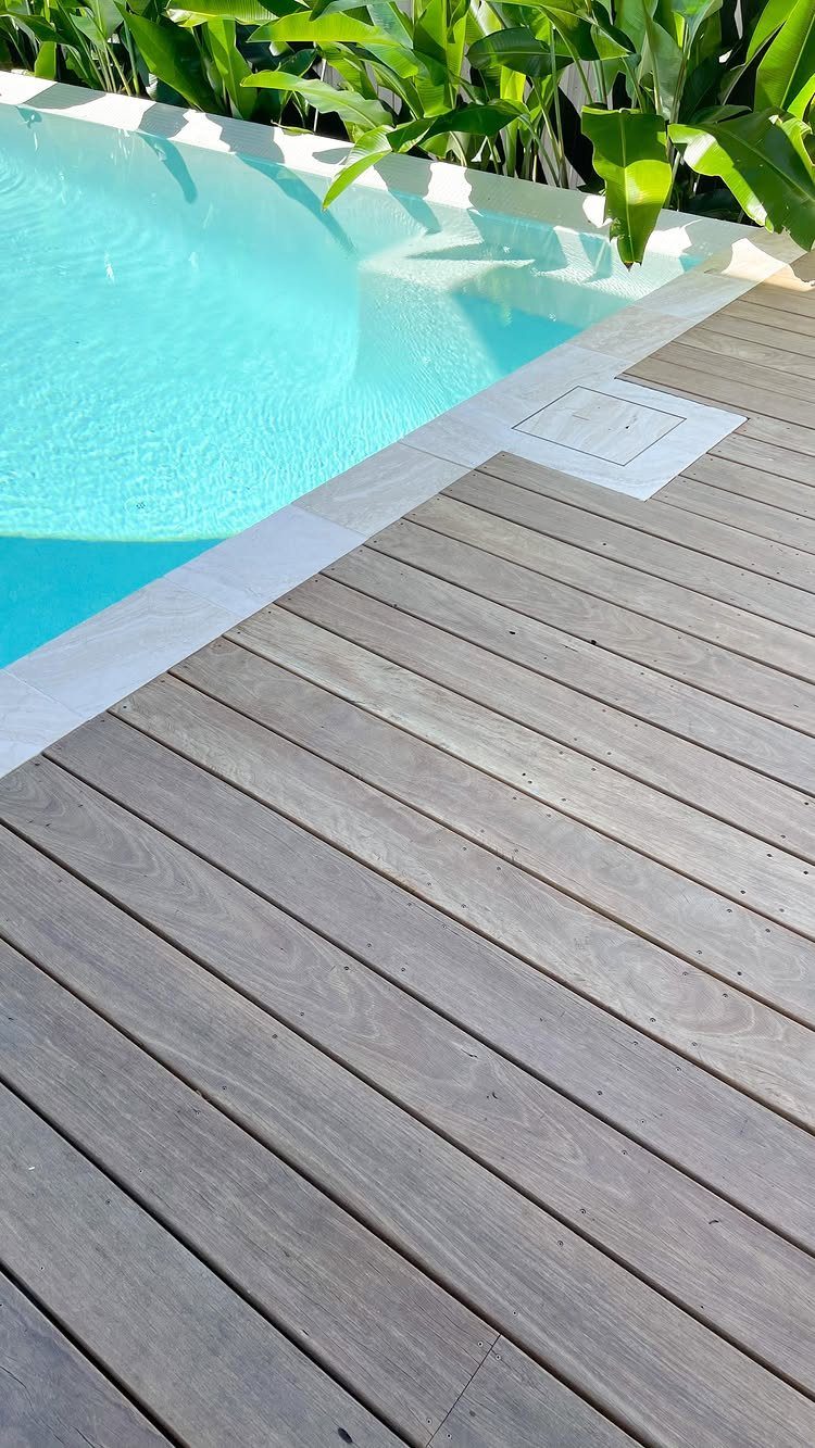 Light-colored wooden deck planks running alongside a bright blue swimming pool with green plants.