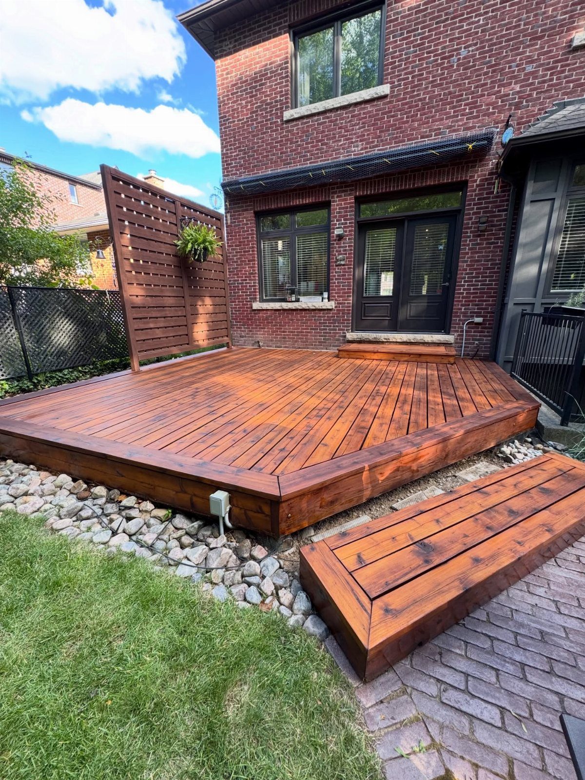 A wooden deck with a reddish-brown stain attached to a brick house next to a lawn.