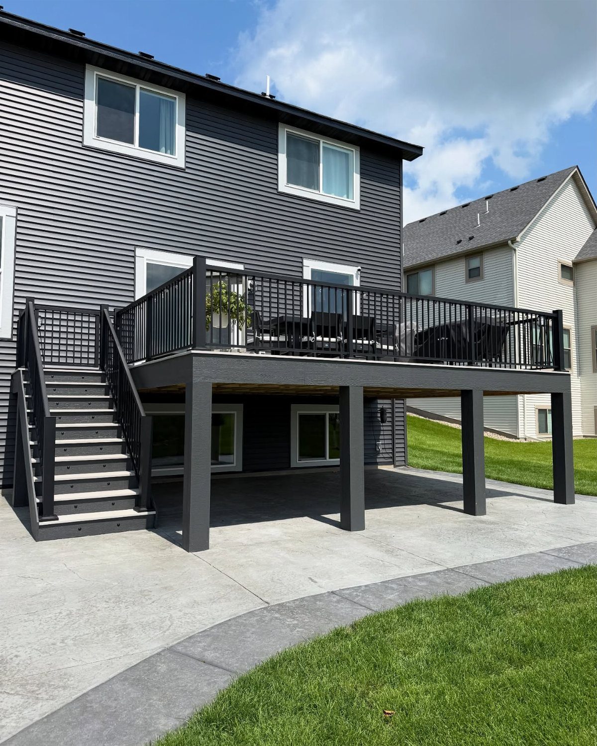 A dark grey wooden deck with black railings overlooking a light grey concrete patio and green lawn.