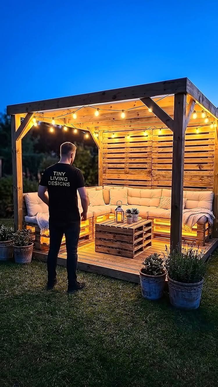 Wooden pallet structure with built-in sofas, a coffee table, and warm string lights in a garden.