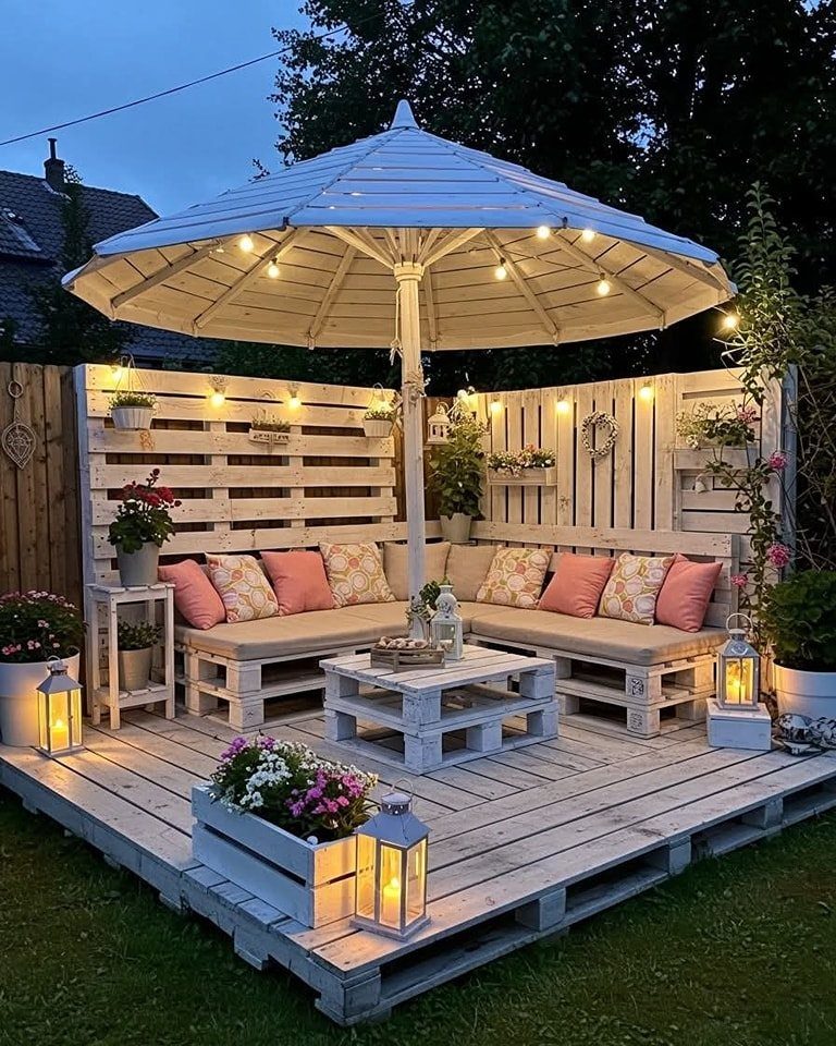 White pallet lounge furniture with pink cushions under a lit patio umbrella on a pallet deck.