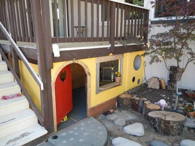 Yellow children's playhouse built under a wooden deck with a red door and stump seating.