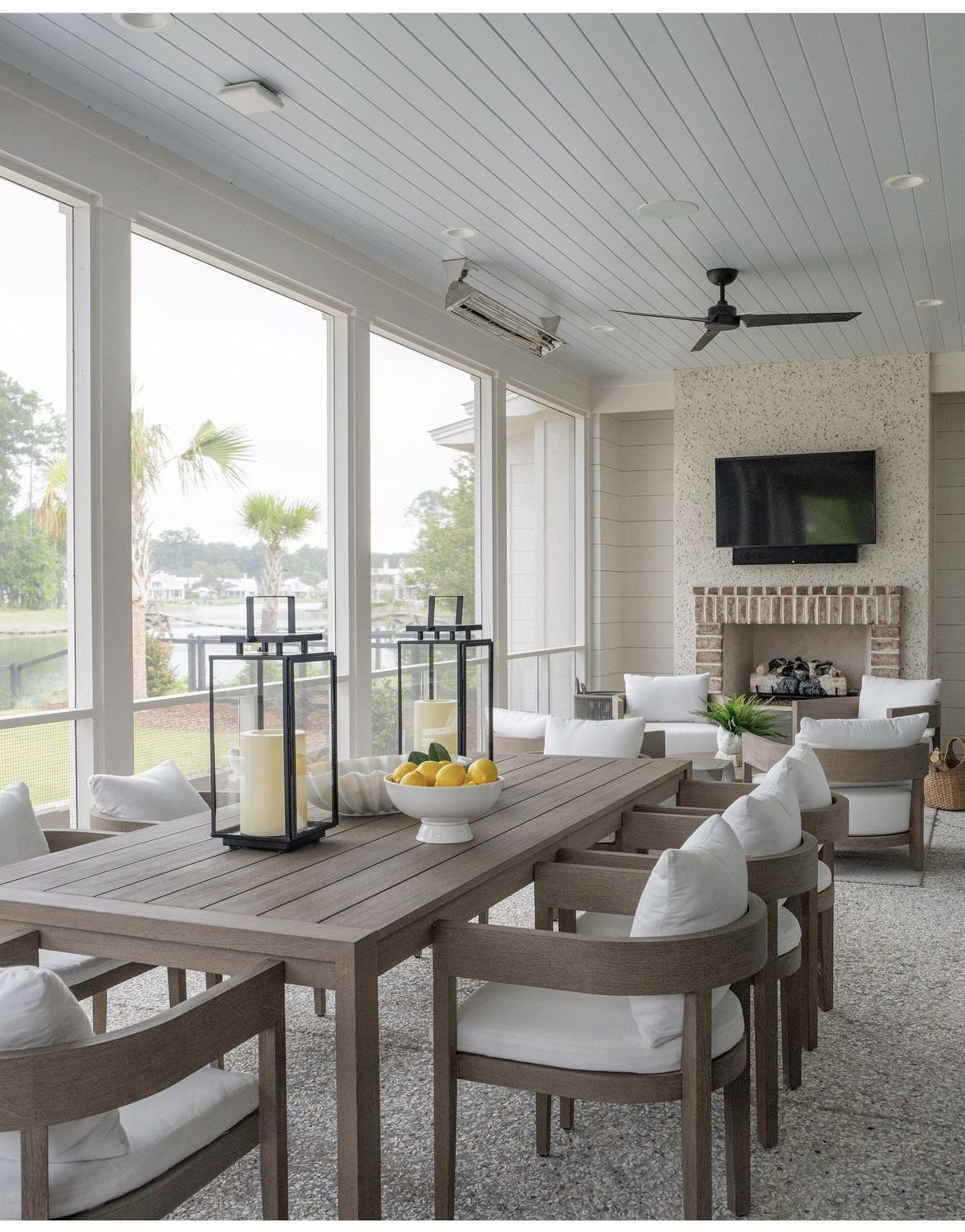 Wooden dining table with white chairs in a bright enclosed outdoor room with fireplace.