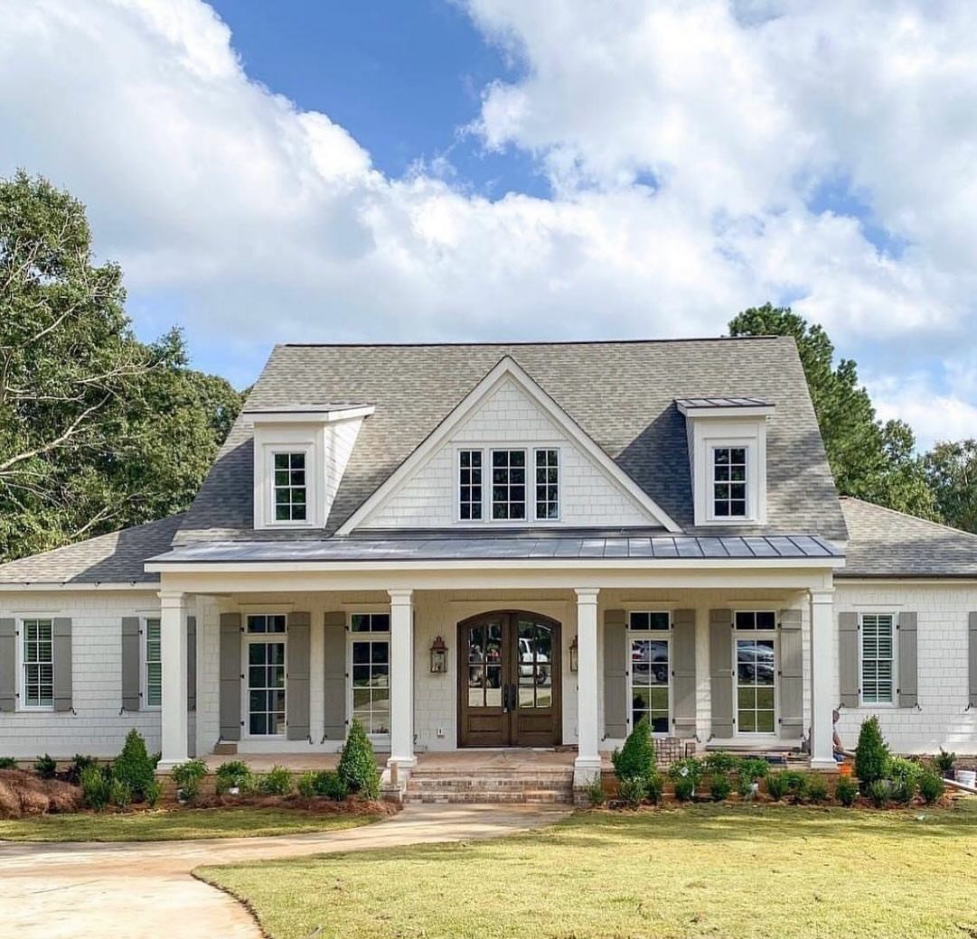 White farmhouse with grey shutters, a large front porch, and a wooden double door under a blue sky.