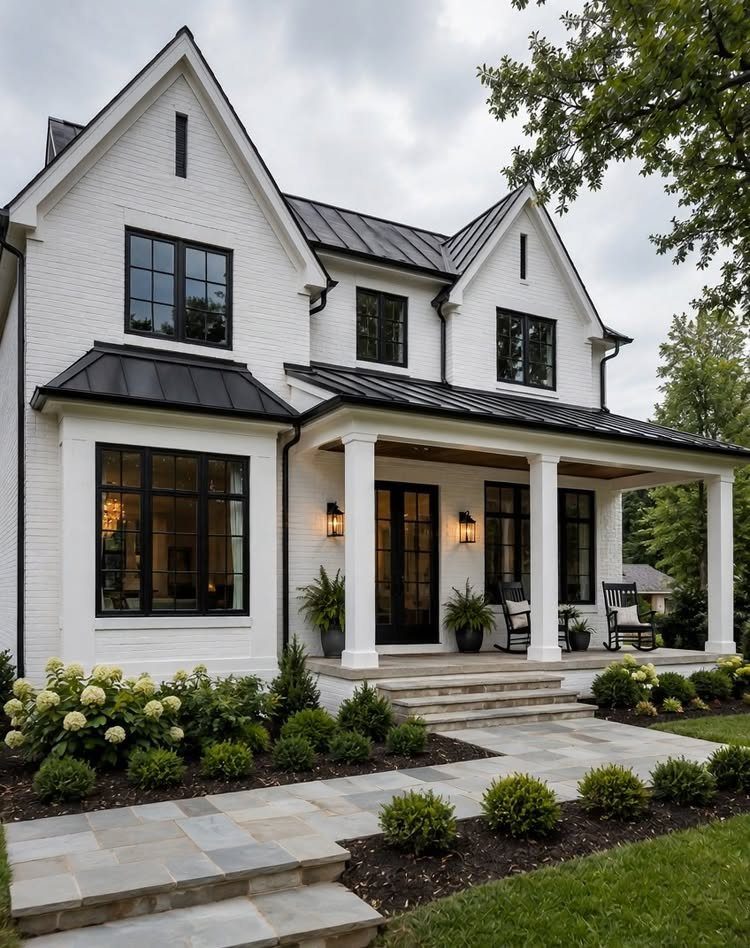 White brick farmhouse with black metal roofing, large windows, and prominent steep gable rooflines.