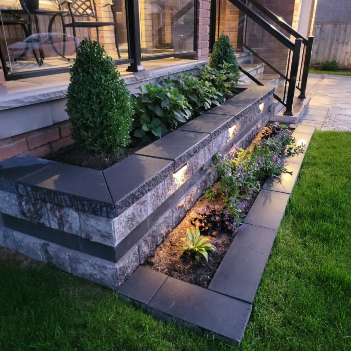 Tiered stone garden beds with green plants and purple flowers in front of a house.