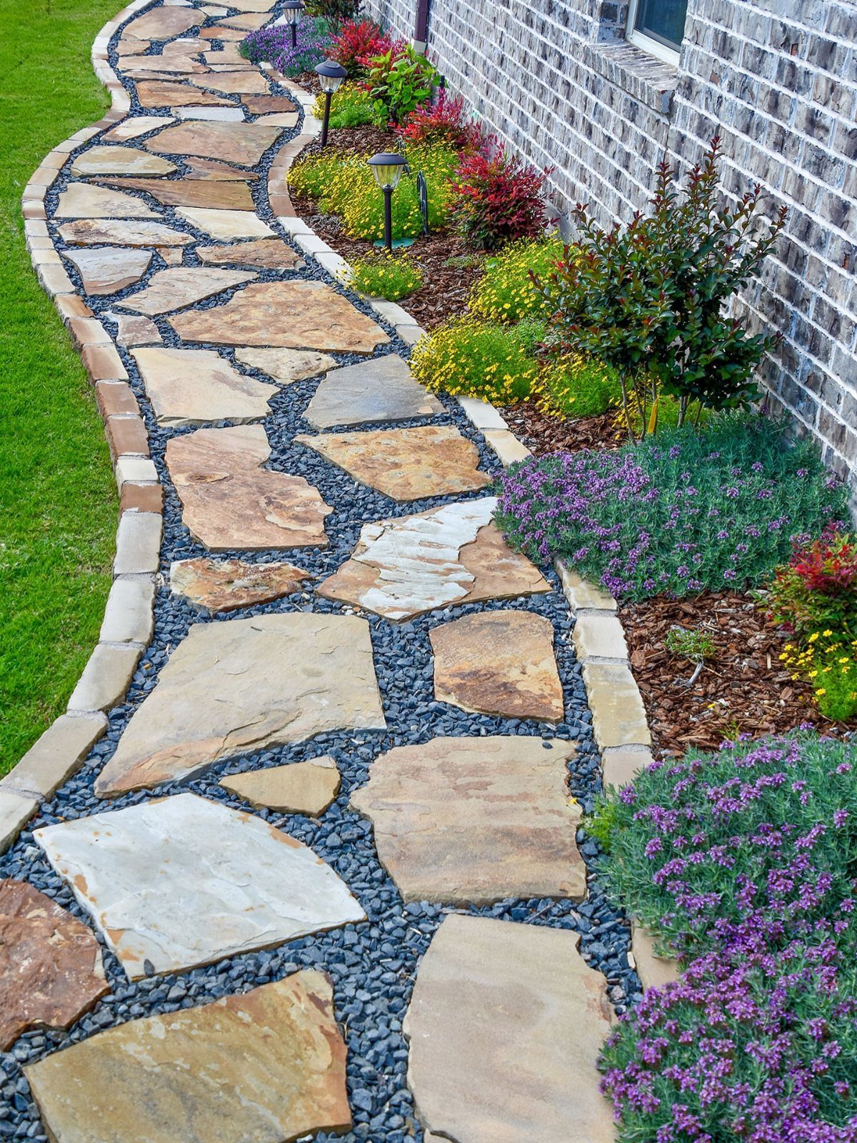 Irregular flagstone pieces set in dark gravel forming a curved walkway alongside a garden and brick wall.
