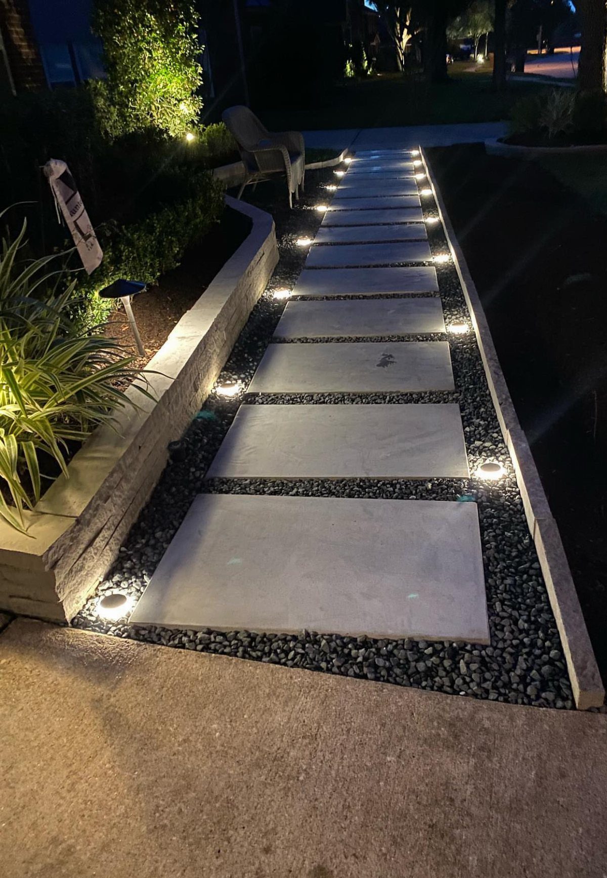 Rectangular concrete pavers create a lit walkway through dark pebbles in a landscaped front yard.