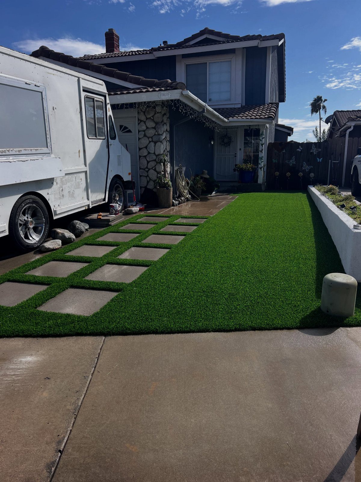 Square concrete pavers arranged in a grid pattern within green artificial turf in a residential front yard.