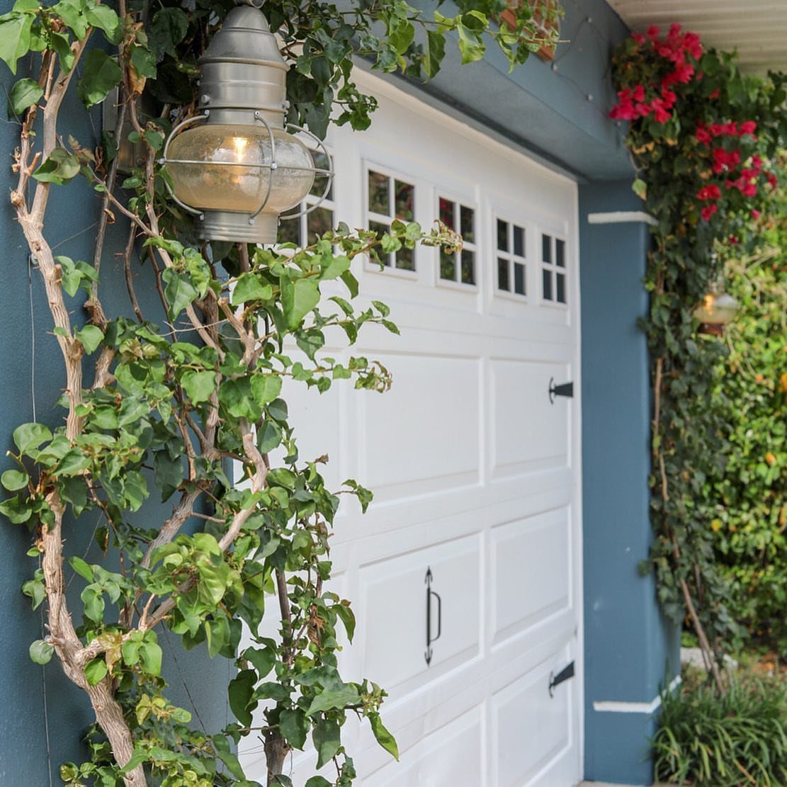 9 Garage Door Makeover Ideas for Instant Curb Appeal 9 White paneled garage door surrounded by green climbing vines and pink flowers against a blue wall.