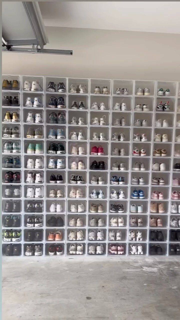 Clear plastic storage boxes filled with many pairs of colorful sneakers stacked against a garage wall.
