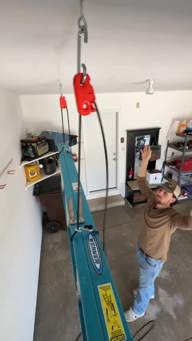 11 Garage Storage Inspiration and Ceiling Systems That Actually Fit 4 A man uses a red pulley hoist to lift a blue ladder in a garage.