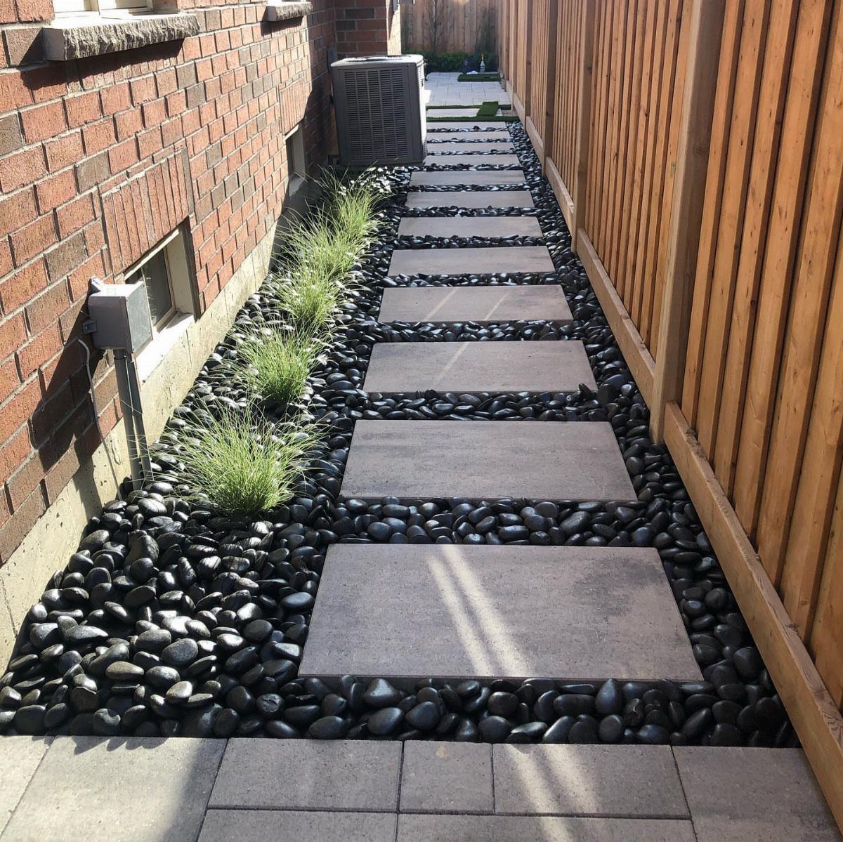 Large rectangular grey concrete pavers set in black river stones along a narrow side yard walkway.