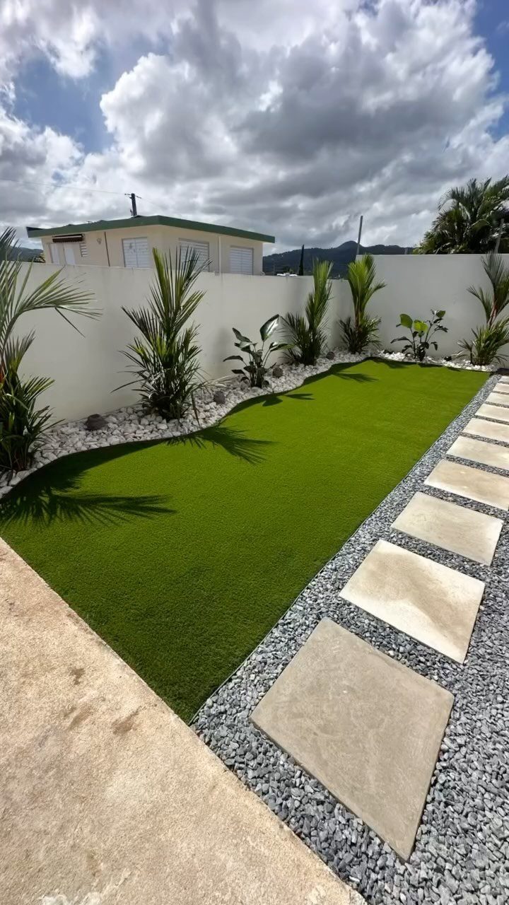 13 Landscaping Plans Layout Ideas to Draw From 13 Artificial turf lawn with tropical plants, white stone borders, and concrete paver walkway in a backyard.