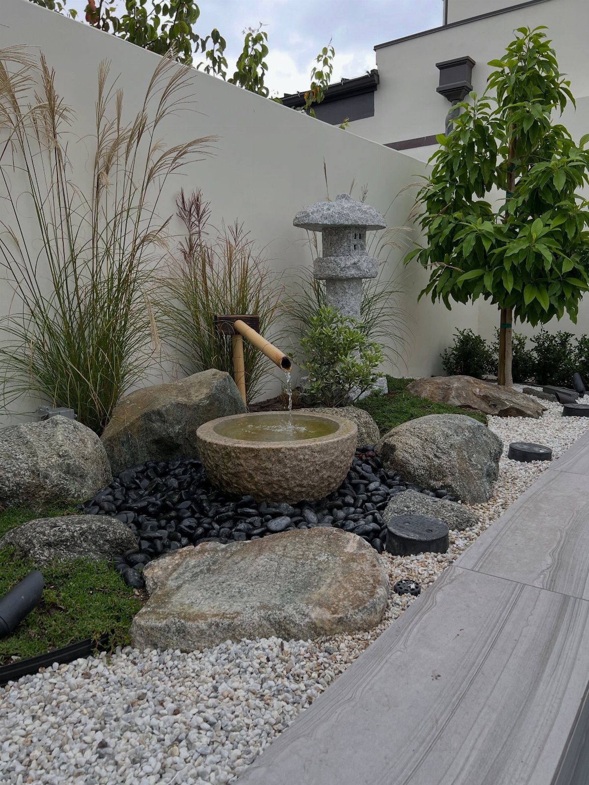 18 Large Yard Ideas for Patios, Pools, Outdoor Kitchens & More 7 A stone water basin with a bamboo spout sits among rocks and pebbles in a Zen garden.