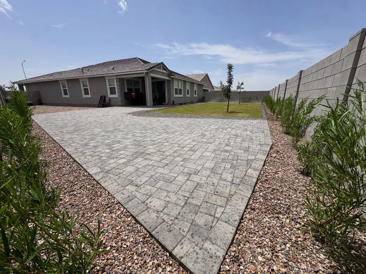 18 Large Yard Ideas for Patios, Pools, Outdoor Kitchens & More 1 Large gray stone paver patio surrounded by gravel and green shrubs in a backyard.