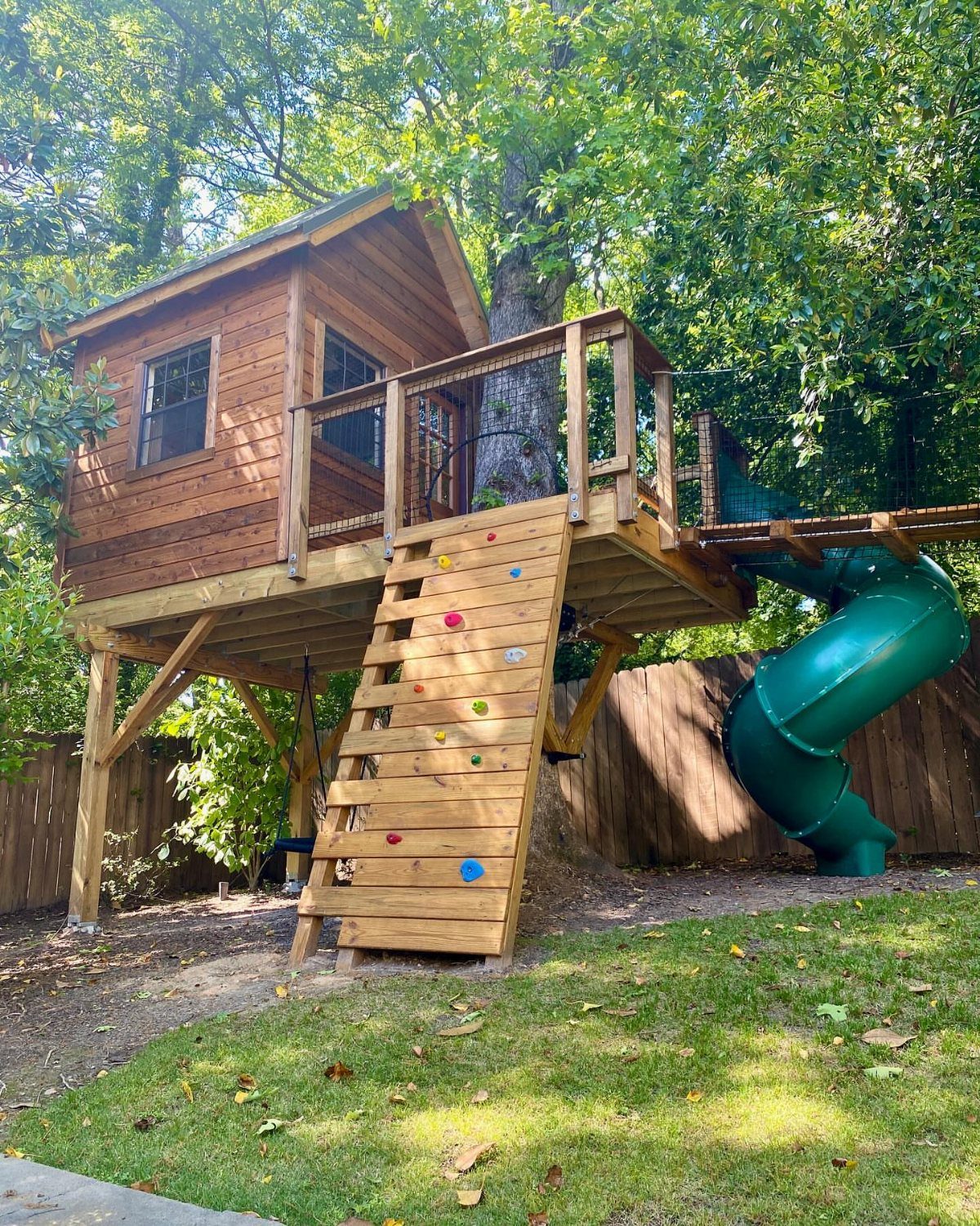 18 Large Yard Ideas for Patios, Pools, Outdoor Kitchens & More 12 A wooden elevated playhouse with a climbing wall and a green spiral slide in a yard.