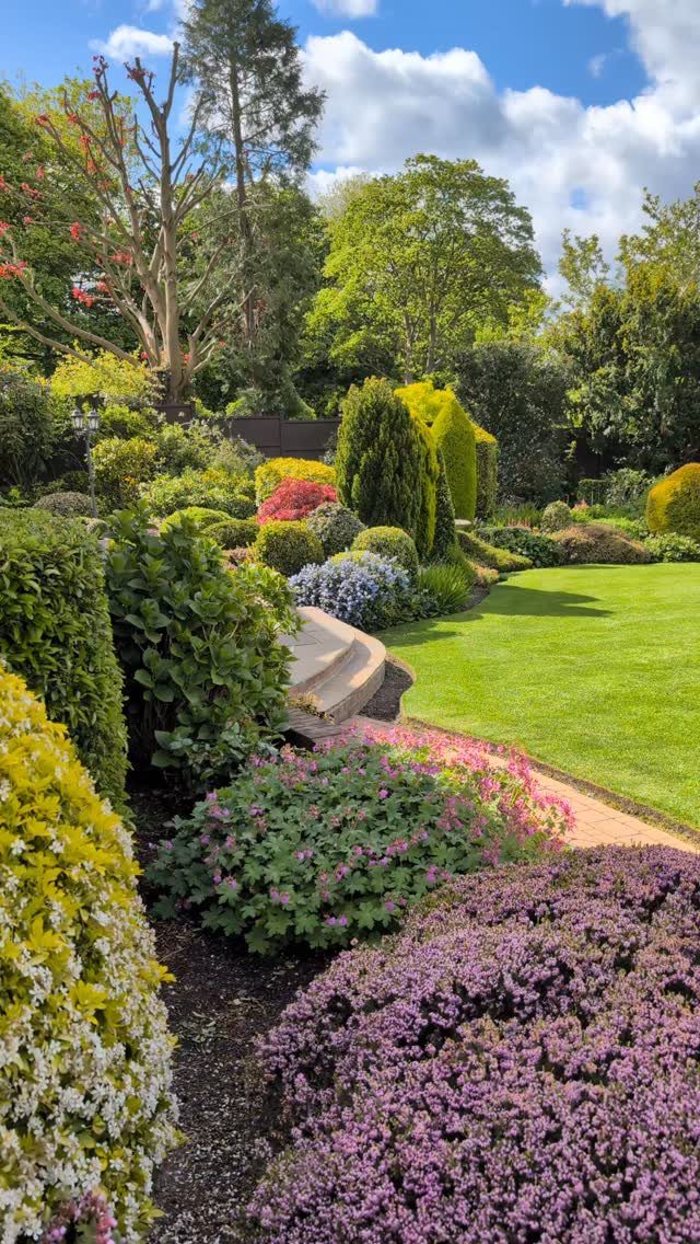 18 Large Yard Ideas for Patios, Pools, Outdoor Kitchens & More 6 Lush garden landscape with diverse flowering shrubs, green lawn, and tall trees under a blue sky.