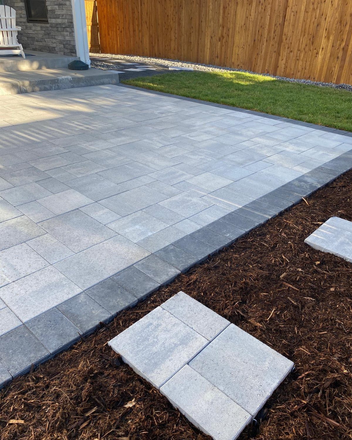 12 Paver Patio Ideas for Fire Pits, Poolside Lounges & Outdoor Rooms 10 Light grey stone paver patio with a dark border next to a lawn and wooden fence.