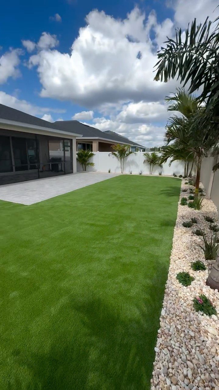 12 Paver Patio Ideas for Fire Pits, Poolside Lounges & Outdoor Rooms 12 Green lawn meeting a light grey paved patio area in a backyard with palm trees.