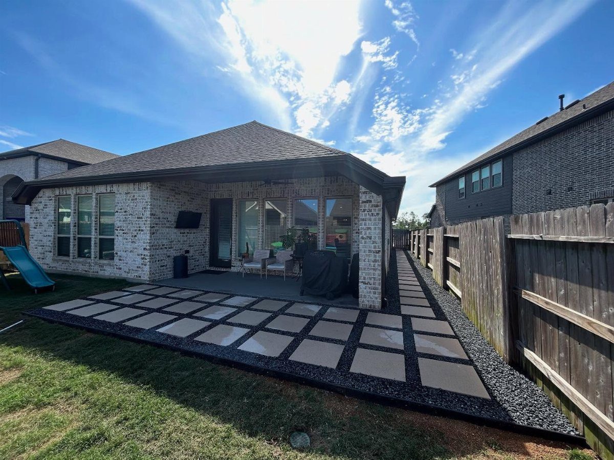 12 Paver Patio Ideas for Fire Pits, Poolside Lounges & Outdoor Rooms 9 Large square paver tiles with dark gravel inlays extending from a brick house toward a lawn.