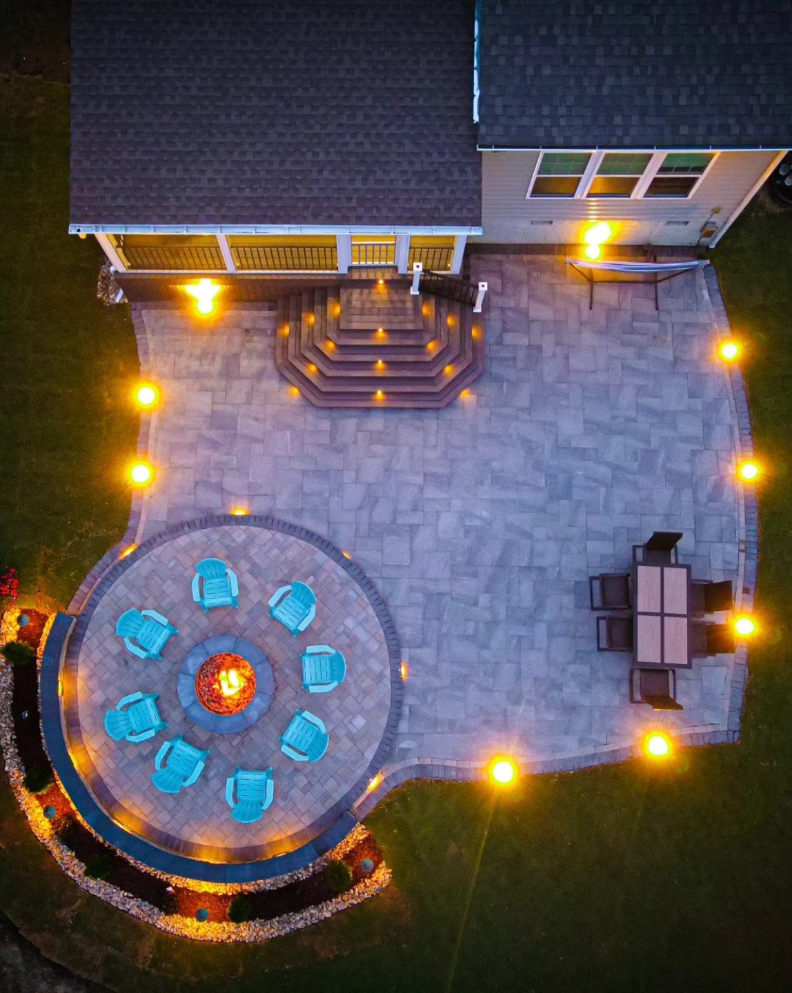 12 Paver Patio Ideas for Fire Pits, Poolside Lounges & Outdoor Rooms 1 Aerial view of a lit stone paver patio featuring a circular fire pit with blue chairs.