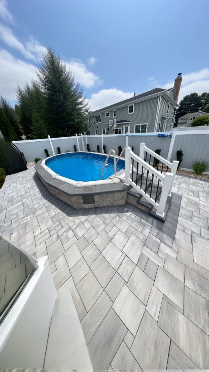 12 Paver Patio Ideas for Fire Pits, Poolside Lounges & Outdoor Rooms 6 A blue swimming pool surrounded by a large grey stone paver patio under a sunny sky.