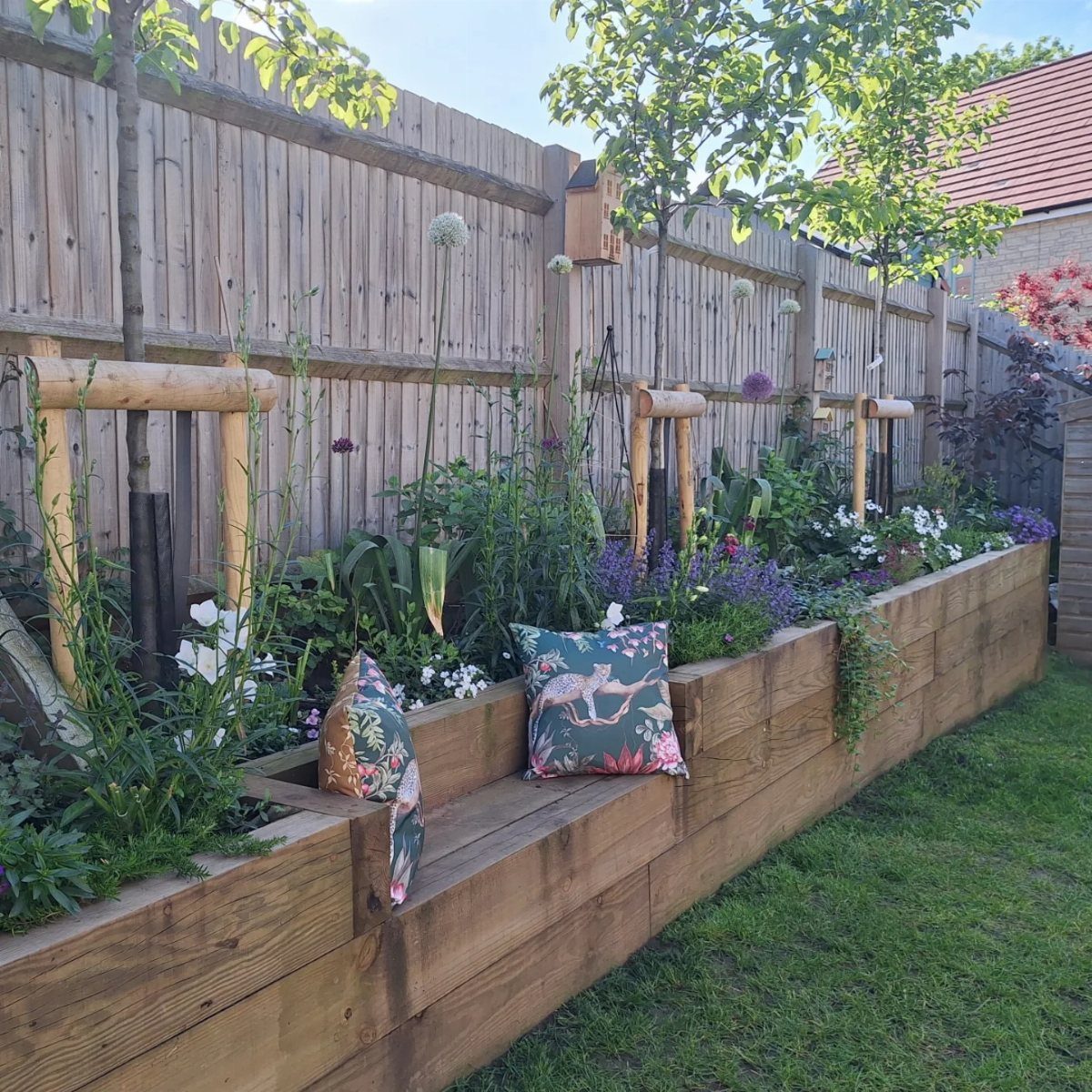 Wooden raised garden bed filled with lush greenery and flowers, featuring decorative pillows on the ledge.